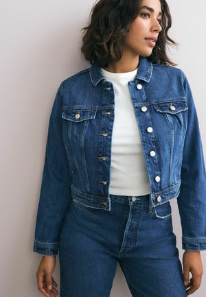 Veste en denim avec un design ajusté, de couleur bleu clair, deux poches poitrine et des finitions en métal argenté, associée à un top blanc côtelé et un jean.