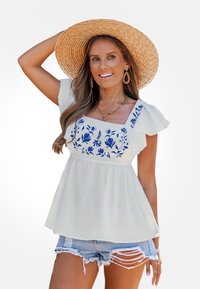 Top blanco con bordado floral azul, mangas cortas con volantes y escote cuadrado, combinado con pantalones cortos de mezclilla desgastados y un sombrero de paja.
