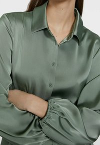 Chemise en satin vert avec col classique, boutonnière à l'avant et manches froncées. Le tissu a une texture lisse et un subtil éclat.