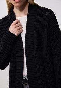 Schwarze gerippte Strickjacke mit Schalkragen, offener Vorderseite und langen Ärmeln, getragen über einem weißen Oberteil; zeichnet sich durch stoffliche Textur und sichtbare Nähte aus.