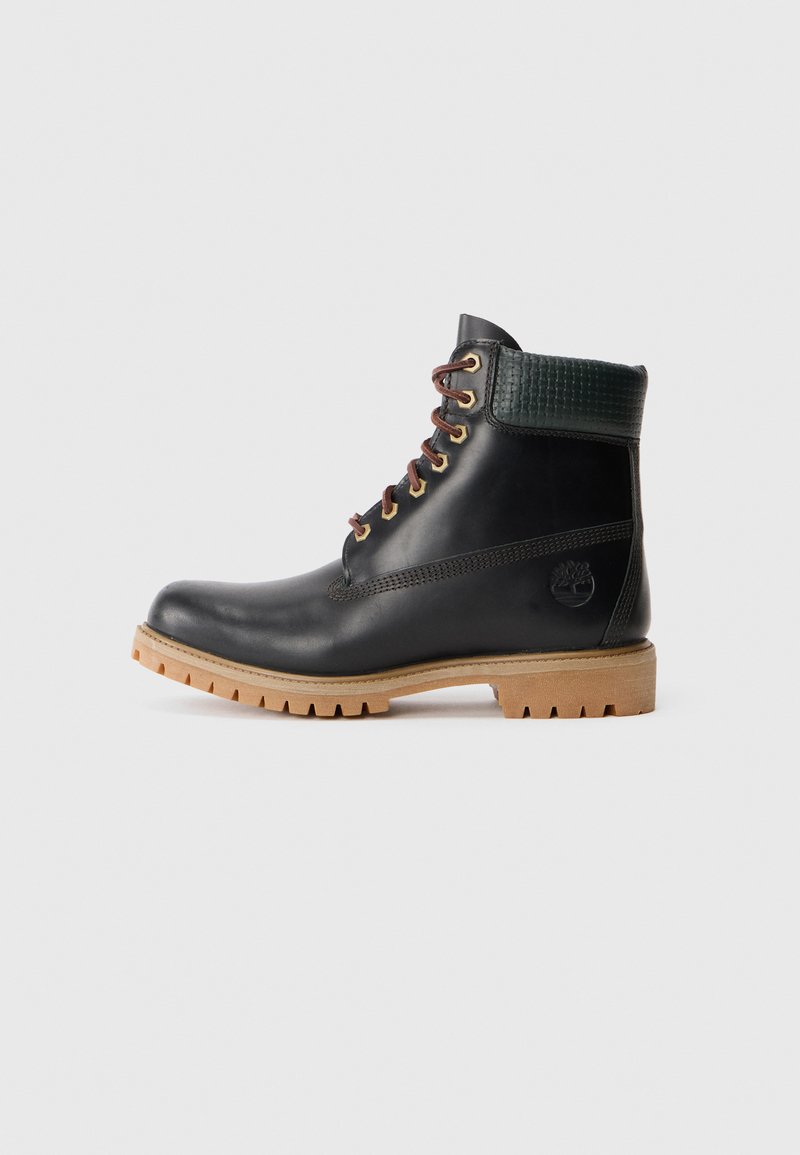 Stivale alla caviglia in pelle nera con lacci marroni, colletto imbottito verde e suola in gomma beige con scanalature profonde, vista laterale su sfondo bianco.