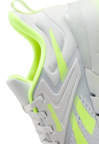 Zapatilla deportiva blanca y amarilla neón con parte superior de malla, diseño elegante, patrones de superposición únicos y una suela texturizada con cordones visibles.