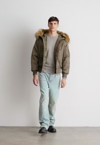 Młody mężczyzna spacerujący w pomieszczeniu, ubrany w brązową kurtkę z futrzanym kapturem, szary sweter, jasnoniebieskie dżinsy i czarne buty na jednolitym tle.