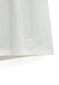 Camiseta blanca de manga corta con una textura suave y lisa y un dobladillo cosido, que presenta un sutil parche de logo cuadrado que dice "POLO CLUB."