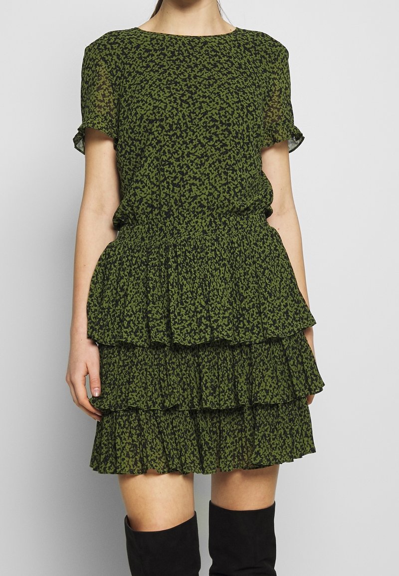 Femme portant une robe à volants à motifs verts et noirs, manches courtes, et des bottes noires montantes, debout devant un fond uni.