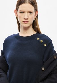 Μπλέ μπλε πλεκτό πουλόβερ με στρογγυλό λαιμόκοψη, που διαθέτει τρία χρυσά κουμπιά στον ώμο. Λεία υφή με εφαρμοστή γραμμή.