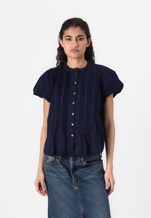 Blusa corta blu navy con colletto arricciato, caratterizzata da pieghe verticali e chiusura con bottoni, abbinata a una gonna di jeans.