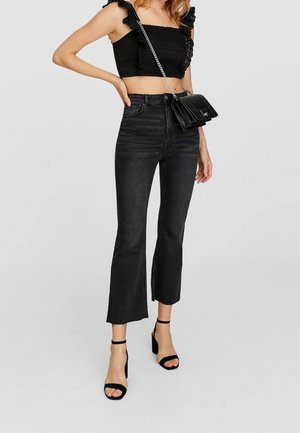 Femme portant un crop top noir à volants, un jean flare noir cropped taille haute, des sandales à talons noires et un sac à main noir.