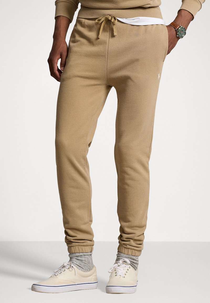 Beige Jogginghosen mit elastischem Bund und Kordelzug, mit einem kleinen Logo an der Seite. Kombiniert mit hellen Sneakers und grauen Socken.