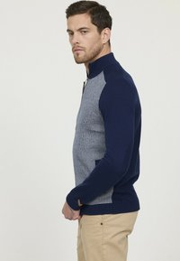 Pull zippé marine et gris avec un col haut, un corps texturé, des manches côtelées et des poches latérales. Conçu avec des couleurs et des matériaux contrastés.