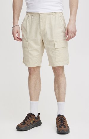 Beige cargo shorts with a relaxed fit, featuring multiple side pockets, made of a lightweight fabric. Worn with brown athletic shoes and white socks.