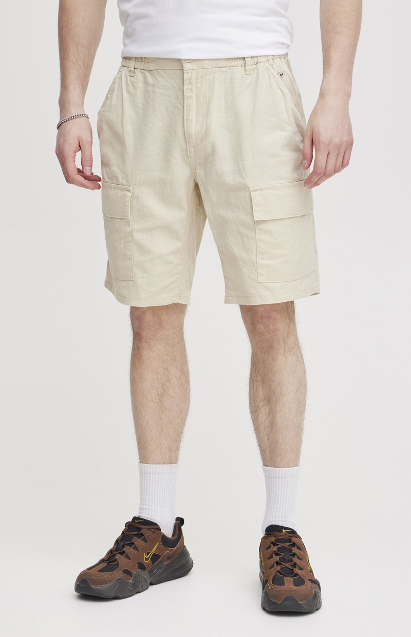 Shorts cargo beiges avec une coupe décontractée, dotés de plusieurs poches latérales, fabriqués dans un tissu léger. Portés avec des chaussures de sport marron et des chaussettes blanches.