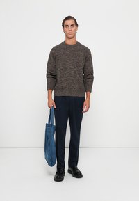Brown textured sweater, navy trousers, and black shoes. Holding a blue denim tote bag. Simple, fitted design with a crew neckline.