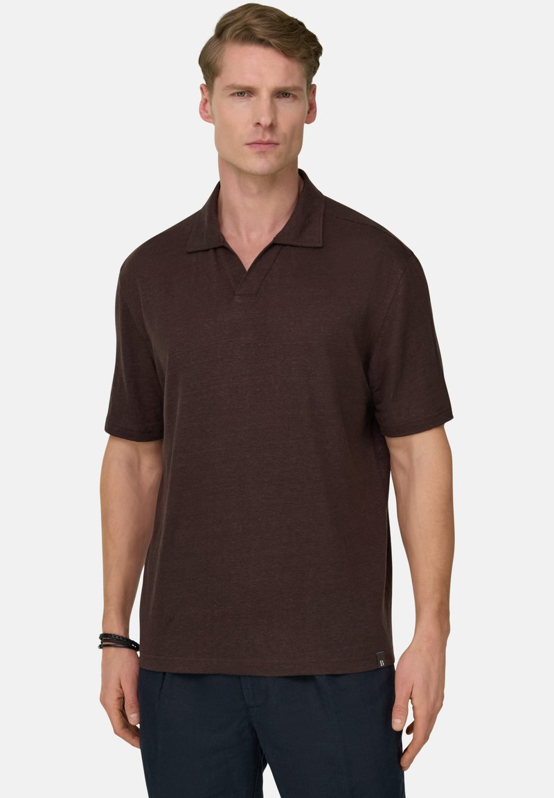 Homme portant un polo à manches courtes marron foncé et un pantalon bleu marine, debout avec une expression neutre sur un fond clair.