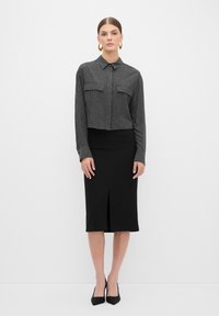 Zwarte cropped shirt met verticale grijze strepen, twee borstzakken, gecombineerd met een zwarte kokerrok met een split aan de voorkant, en zwarte hoge hakken.