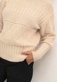 Beige gestrickter Pullover mit strukturierten Rautenmuster, geripptem Saum und Bündchen sowie einem entspannten Schnitt. Kombiniert mit schwarzen Hosen.