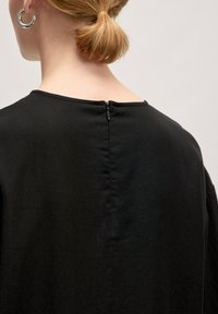 Zwarte kleding met een ronde halslijn, ritssluiting op de rug en een gladde textuur. De stof valt zacht om de schouders.