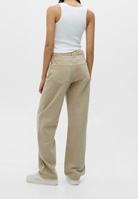 Jeans beiges taille haute avec une coupe décontractée, dotés de poches arrière et de coutures visibles. Associés à un débardeur blanc et des baskets blanches.