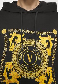 Černá mikina s kapucí s velkým zlatým barokním vzorem a názvem značky "Versace Jeans Couture" ve stylizovaném textu.