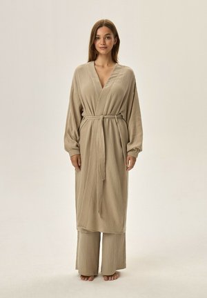 Young woman standing barefoot in beige textured long robe tied at the waist with matching wide-leg pants against a plain background.