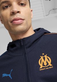 Felpa con cerniera di un blu navy scuro, con logo e testo arancione del Marsiglia, caratterizzata da dettagli azzurri leggeri sulle spalle. Tessuto morbido.