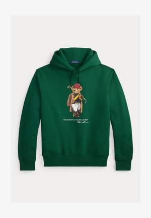 Hanorac verde din bumbac, cu un desen grafic cu un urs purtând o haină maro și o pălărie roșie, ținând o croșetă de golf, și pantaloni albi. Sigla semnăturii sub imagine.