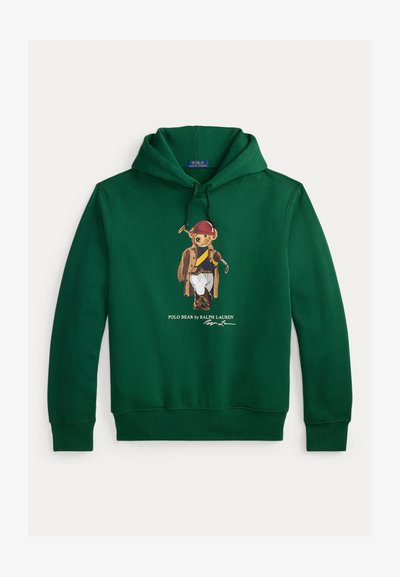 Zelena pamučna majica s kapuljačom s grafikom medvjeda u smeđem kaputu s crvenom kapom, koji drži golf palicu i nosi bijele hlače. Ispod je prepoznatljiv logo.