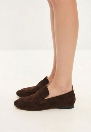 Person iført mørkebrune ruskind slip-on loafers med lave sorte hæle, stående på en lysefarvet overflade.