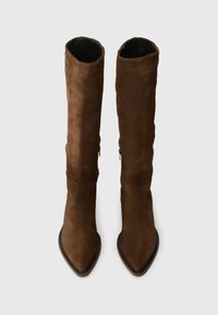 Barna velúr térd fölé érő csizmák, hegyes orral, letisztult dizájnnal és oldalsó cipzárral a könnyű felvétel érdekében. Simaság és matt felület jellemzi.