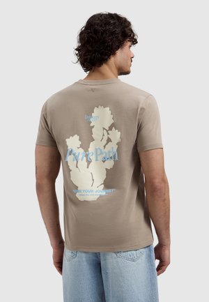 Man met krullend haar, van achteren gezien, draagt een beige t-shirt met witte bloemenprint en blauwe tekst "Pure Path" en lichtblauwe spijkerbroek.