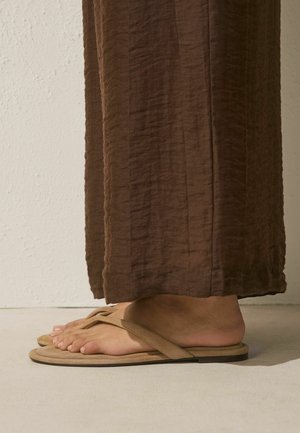 Person wearing beige flat sandals with thong straps and loose brown textured pants standing on a light floor against a white wall.
