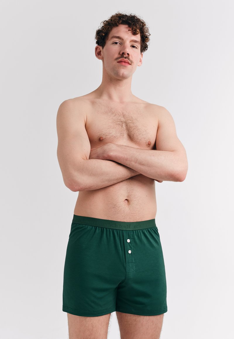 Grüne Baumwollboxershorts mit Knopfverschluss, elastischem Bund mit Logo und glatter Textur. Das Model posiert mit verschränkten Armen.