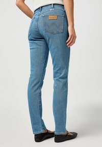 Jeans ajustados de color azul claro, con costuras visibles, diseño de cinco bolsillos y una etiqueta de cuero que dice "Wrangler" en la parte trasera.