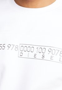 Bílý bavlněný trik s vystouplým stříbrným textovým designem, na kterém jsou zvýrazněna čísla a nápis "DIESEL". Měkká textura, kulatý výstřih.