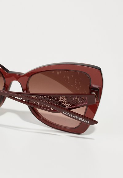 Burgundijas saulesbrilles ar pārmērīgu kaķa acs formu, tonētām lēcu brillēm un sarežģītu mežģīņu raksta tempļiem, kas rotāti ar zīmola logo.