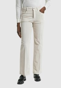 Pantalons flare en côtes de velours blancs avec poches avant, taille haute et surface texturée. Assortis à des mocassins noirs sur un fond neutre.