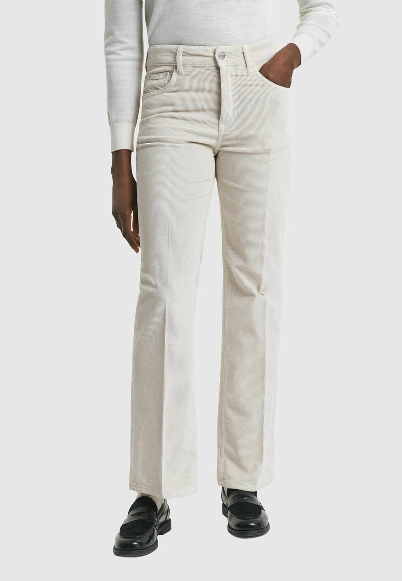 Witte fluweelachtige flared broek met voorzakken, hoge taille en een gestructureerd oppervlak. Gecombineerd met zwarte instappers op een neutrale achtergrond.