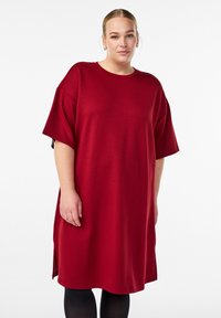 Červené nadrozmerné šaty z jemného materiálu, s krátkymi rukávmi, okrúhlym výstrihom a bočnými štrbinami pre väčšiu voľnosť pohybu.