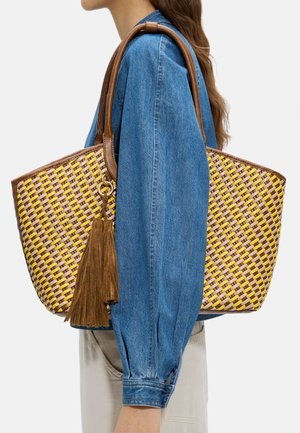 Kobieta w niebieskiej kurtce jeansowej trzyma dużą żółto-brązową plecioną torbę na ramię z skórzanymi uchwytami i brązowymi frędzlami.