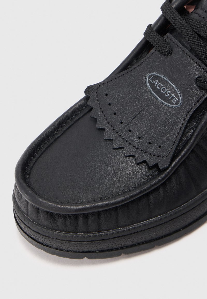 Sort læder Lacoste loafers med teksturerede kanter, dekorative perforeringer og tyk gummisål på hvid baggrund.