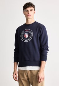 Granatowy nadruk z okrągłym logo, na którym widnieją czerwone i białe paski, tekst „GANT 1949” oraz trzy gwiazdki; ściągacze przy mankietach i dole.