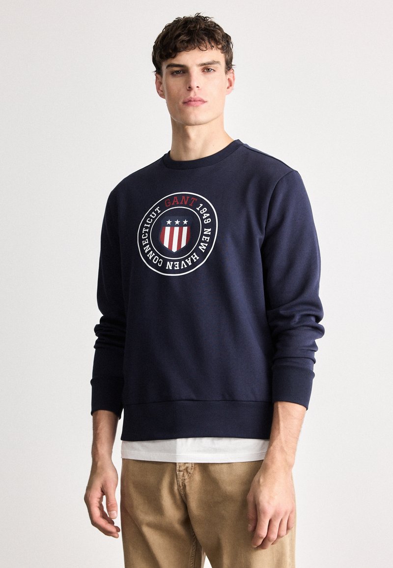 Granatowy nadruk z okrągłym logo, na którym widnieją czerwone i białe paski, tekst „GANT 1949” oraz trzy gwiazdki; ściągacze przy mankietach i dole.