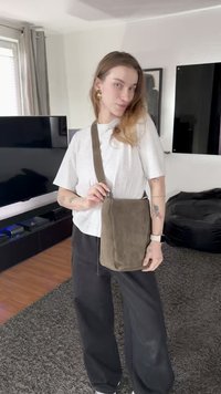 Brūna zamšādas plecu soma ar garu siksnu, kvadrātveida forma, gluda tekstūra un minimālistisks dizains, ko nēsā cilvēks ikdienišķā apģērbā.