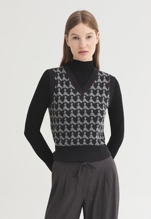 Femme portant un top noir à manches longues sous un pull sans manches noir et gris à motifs, avec un pantalon gris foncé à cordon.