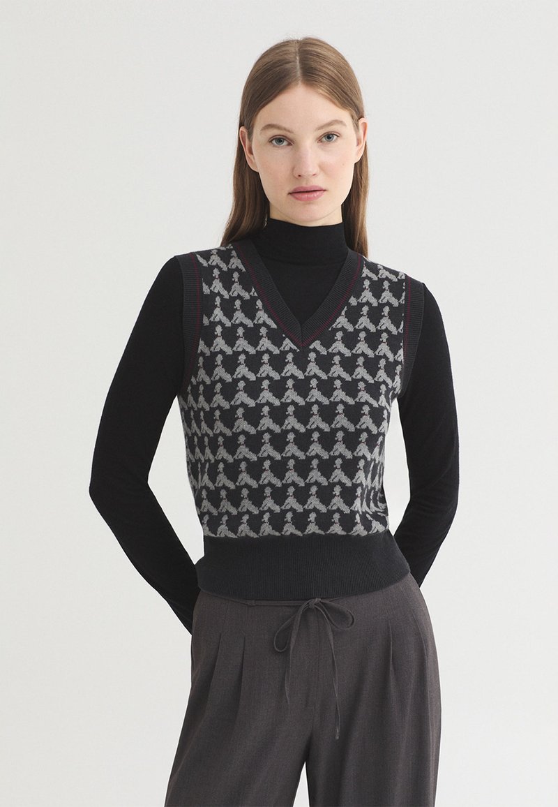 Femme portant un top noir à manches longues sous un pull sans manches noir et gris à motifs, avec un pantalon gris foncé à cordon.