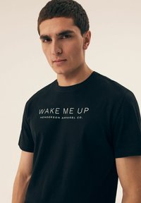 Czarna bawełniana koszulka z białym napisem "WAKE ME UP" oraz "HENDERSON APPAREL CO." nadrukowanym z przodu. Okrągły dekolt. Krótkie rękawy.