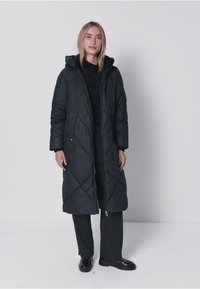 Zwarte gewatteerde lange jas met capuchon, ruitsteekpatroon, zijknopen en halflange snit, gecombineerd met donkere broek en zwarte schoenen.