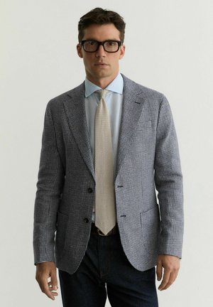 Mężczyzna w szarej kratkowanej marynarce, jasnoniebieskiej koszuli, beżowym dzianinowym krawacie, ciemnych dżinsach i czarnych okularach, stojący na tle jednolitego tła.