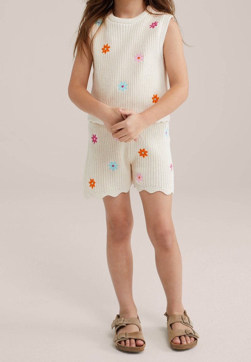 Enfant portant un haut sans manches en maille blanche et un short décoré de fleurs brodées colorées, debout les mains jointes, sandales beiges aux pieds.