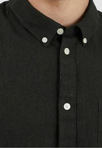 Chemise noire à col, avec des boutons blancs et un tissu texturé. Inclut une poche poitrine sur le côté gauche.